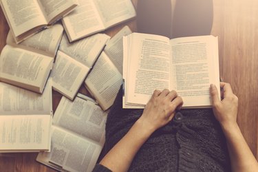 A person reads an open book on their lap, with several other books spread around them.